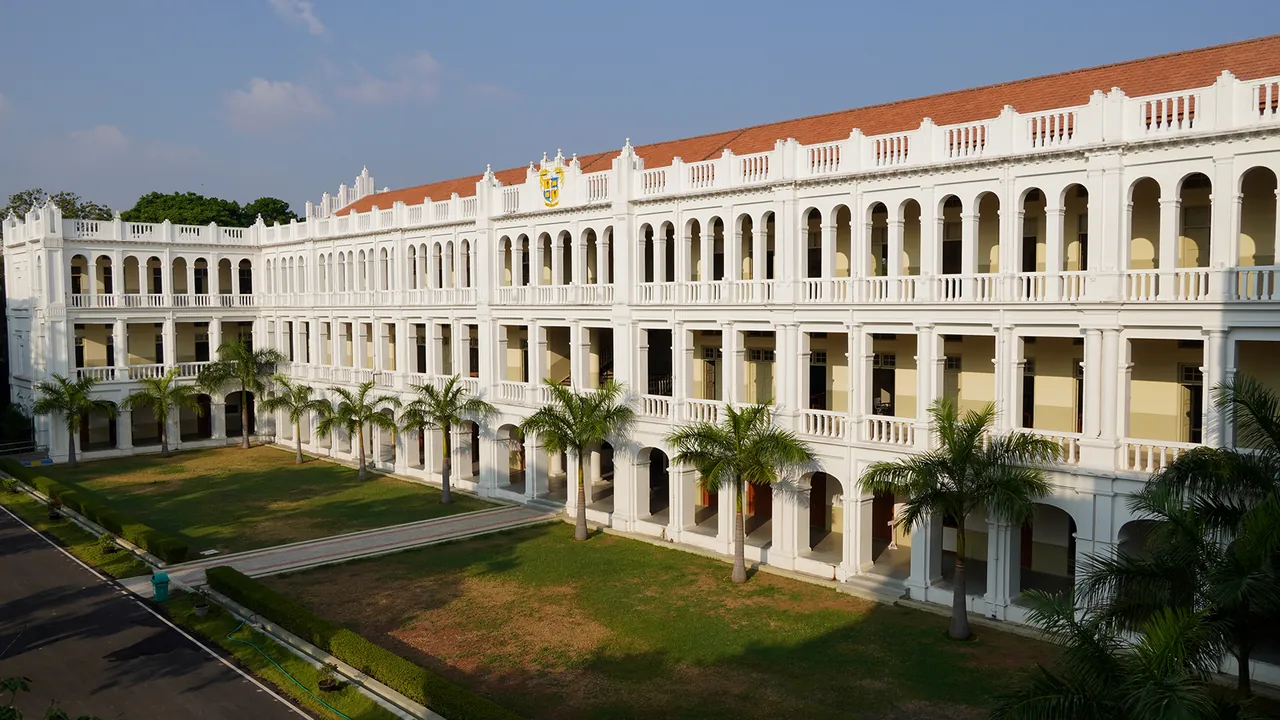 The main building of Loyola College, white with a red tiled roof, seen across its front lawn and palms
