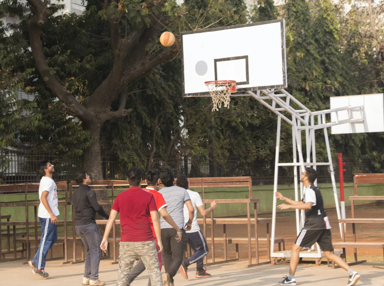 Students playing basketball on the outdoor court of the IES campus, under trees. Shows the grounds, not the building. Hosted on the Webflow CDN the college's site is served from.