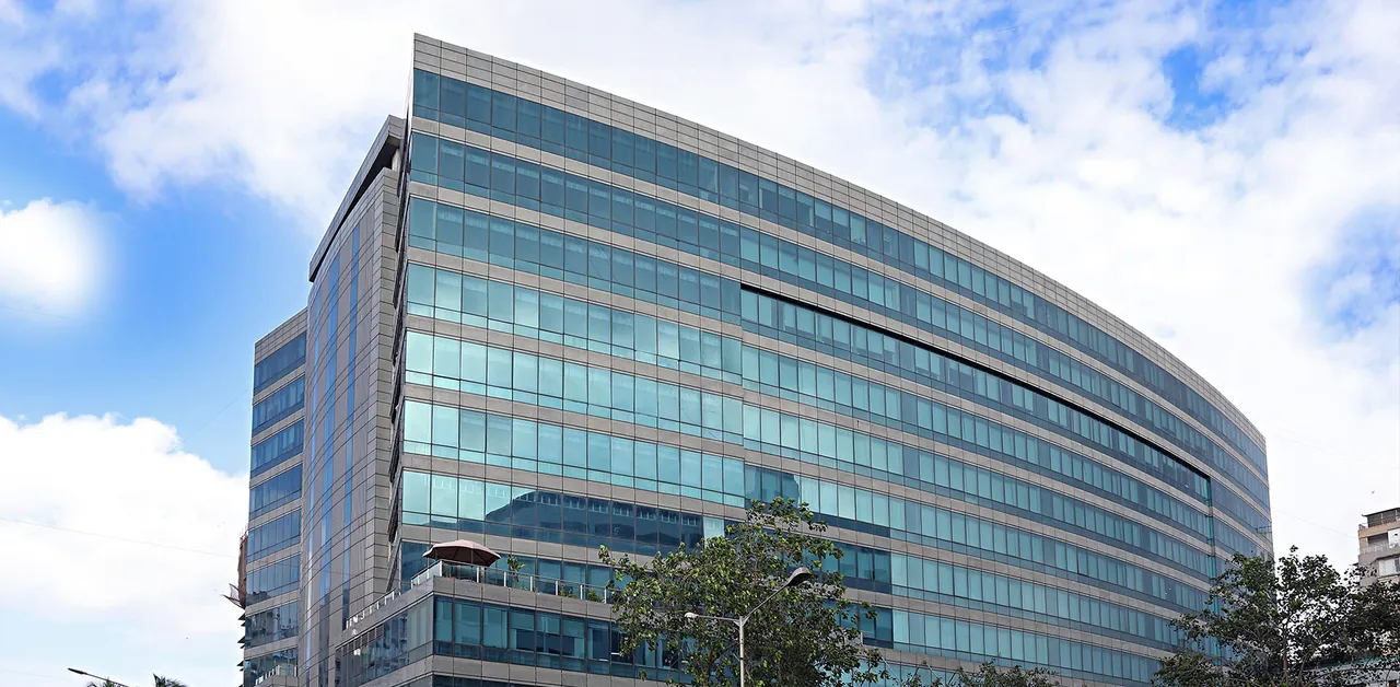 The curved blue-glass NMIMS building from street level, under a cloudy blue sky.