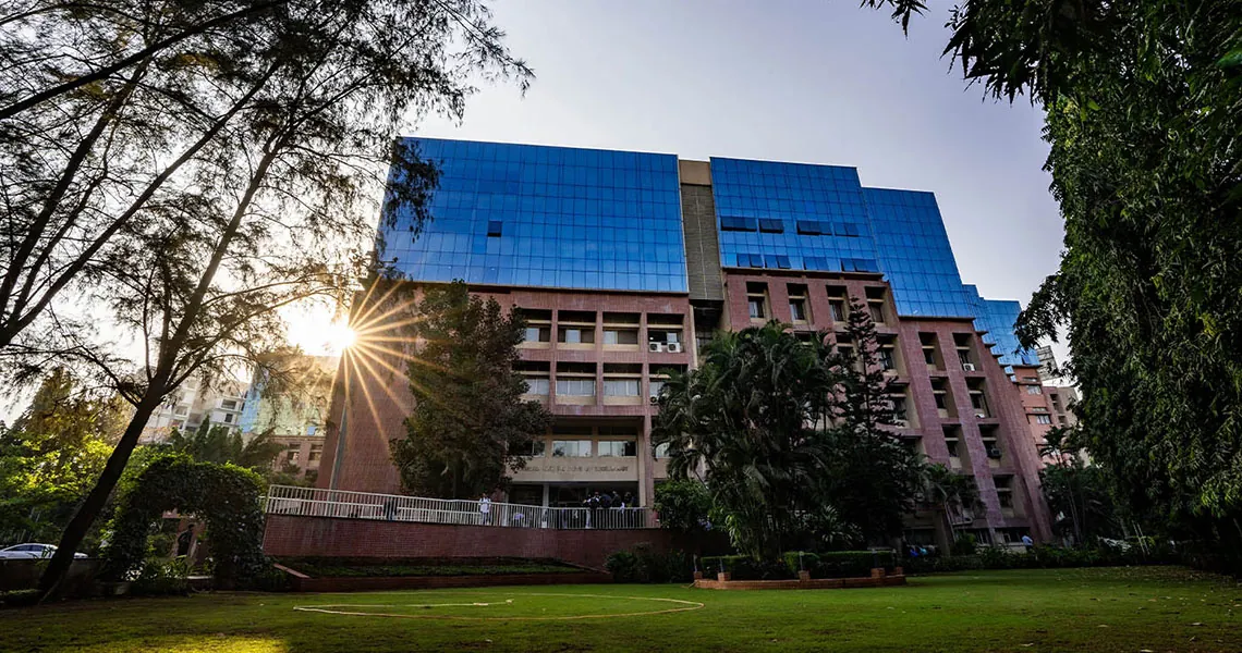The Ramrao Adik Institute of Technology building, pink stone with blue glass upper floors, from the lawn with the sun behind a tree. Hosted on the university's own S3 bucket used by dypatil.edu.