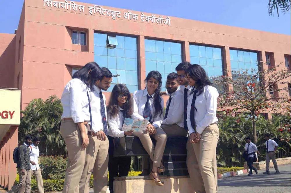 The front of the Symbiosis Institute of Technology building at Lavale, its name in Devanagari across the top, with a group of students seated in the foreground. Weaker than ideal: the students fill about a third of the frame; the cleaner gate photograph on the same page (sit-campus-1.jpg) is only 622 pixels wide